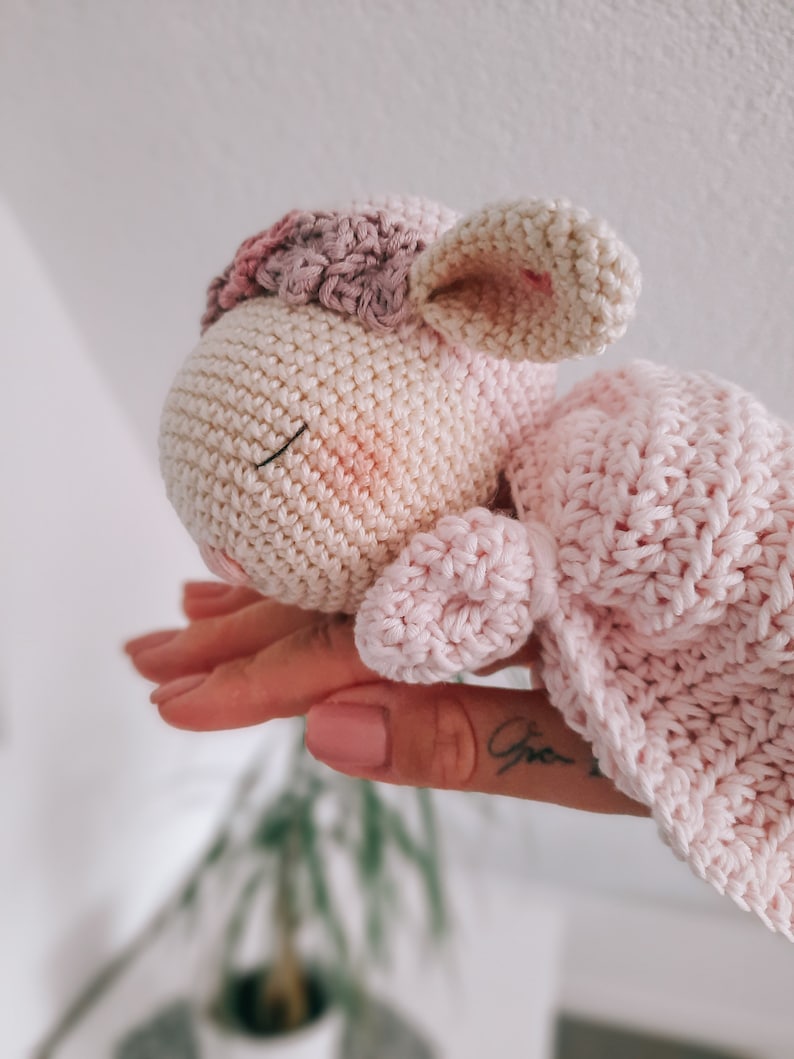 Op de afbeelding: Een gehaakt roze en wit lammetje met een roze deken. Het lammetje heeft een gesloten oog en een kleine mond.