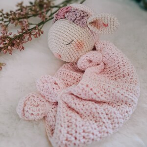 Op de afbeelding: Een gehaakt roze en wit lammetje met een bloemenkrans, gewikkeld in een roze gehaakte deken.