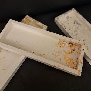 Könnte beinhalten: Drei rechteckige Betontabletts mit Blattgold-Akzenten. Die Tabletts sind weiß mit einer gesprenkelten Oberfläche.