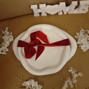 Pode incluir: Bandeja de cerâmica branca com borda ondulada, amarrada com uma fita de veludo vermelho. A bandeja está sobre uma superfície marrom com flores brancas.