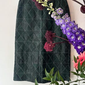 May include: A dark green skirt with a geometric pattern, hanging on a metal hanger. The skirt has a textured appearance and a knee-length cut. The background features purple and pink flowers and green foliage.