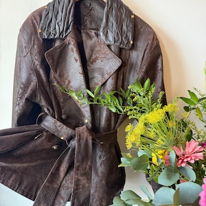 May include: A brown leather jacket with a snakeskin pattern, double-breasted design, and a matching belt. The jacket has a pleated collar and gold-coloured buttons. The jacket is hanging on a wooden hanger.
