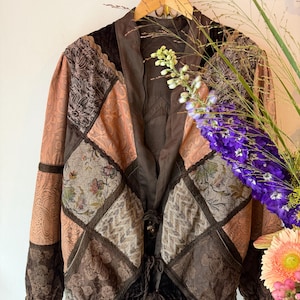 May include: A long-sleeved jacket featuring a patchwork design of brown, tan, and peach fabrics with various floral and geometric patterns. The garment has a V-neckline, a solid brown collar, and two front tie closures with dangling decorative beads.