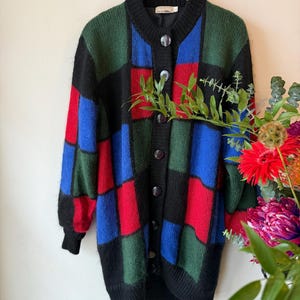 Op de afbeelding: Een lange cardigan met knopen in een patchwork-ontwerp van zwarte, groene, rode en blauwe vierkanten. De trui heeft een zwarte kraag, manchetten en een knoopsluiting. De cardigan hangt aan een houten hanger.