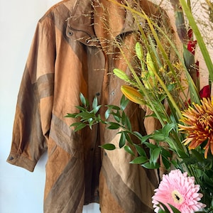 May include: A long-sleeved jacket crafted from panels of tan and brown suede leather. The garment features a high collar, a front button closure, and a drawstring hem. The design consists of alternating diagonal stripes of varying brown tones.