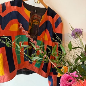 Könnte beinhalten: Eine farbenfrohe Jacke mit Muster und einzigartigem Design. Die Jacke zeigt eine Mischung aus orangefarbenen, marineblauen, gelben und grünen geometrischen Formen. Sie hängt an einem Holzbügel vor einer weißen Wand, mit einem Blumenarrangement im Vordergrund.