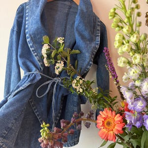 Op de afbeelding: Een blauwe spijkerjas met een brede kraag en een strik in de taille. De jas hangt aan een houten hanger. Een boeket bloemen met oranje, paarse en witte bloemen staat op de voorgrond.
