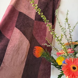 Może przedstawiać: Spódnica w stylu patchwork w odcieniach bordowego, malinowego i różowego wisi na wieszaku. Projekt spódnicy zawiera geometryczne panele. Bukiet kolorowych kwiatów i zieleni znajduje się na pierwszym planie.