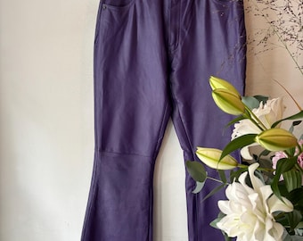 Può includere: Pantaloni in pelle viola con design a zampa d'elefante. I pantaloni sono appesi a una gruccia argentata contro un muro bianco. Presentano un classico design a cinque tasche e una chiusura con bottone e cerniera.