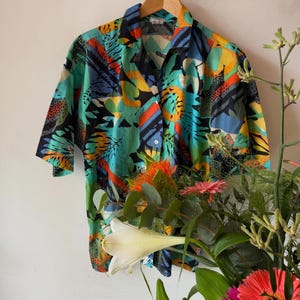 Op de afbeelding: Een shirt met korte mouwen met een levendige tropische print in teal, zwart, oranje, geel en blauw. Het shirt hangt aan een houten hanger tegen een witte muur. Het ontwerp heeft abstracte blad- en geometrische vormen.