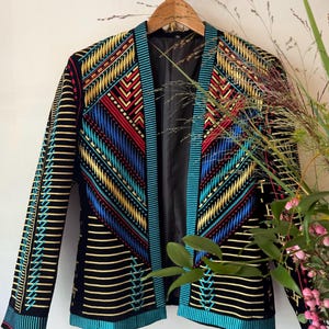 Op de afbeelding: Een zwarte blazer met ingewikkeld borduurwerk in levendige tinten goud, rood en turquoise. Het jasje heeft een gestructureerde open voorkant, lange mouwen en een geometrisch patroon van horizontale lijnen en chevronvormen op de voorpanden.