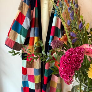 May include: A long, open-front robe constructed from a patchwork of multi-colored corduroy fabric squares. The garment features a solid black trim along the collar and front opening, with a plain cream-colored interior lining visible.