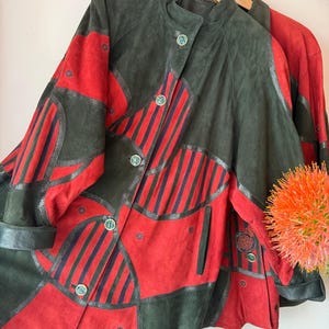 Op de afbeelding: Een vintage suède jas in een patchwork-ontwerp, met rode en donkergroene panelen met zwarte accenten. De jas heeft een ronde kraag, knoopsluitingen en een uniek gestreept patroon. De mouwen zijn omgeslagen.