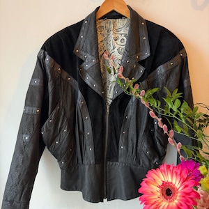 May include: Black leather jacket with suede panels and silver stud embellishments. The jacket has a zip closure, a notched collar, and a distinctive design. The lining has a patterned design. A vintage-style garment.