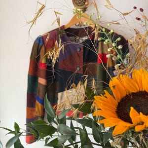 Op de afbeelding: Een jas met lange mouwen voorzien van een veelkleurig patchworkontwerp met geometrische panelen in tinten rood, blauw, groen, bruin en zwart. De jas heeft een ronde halslijn, twee donkere knopen aan de voorzijde en een gevoerde binnenzijde met patroon.