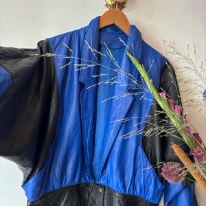 May include: A vintage leather jacket with a bold colour-block design. The jacket has a vibrant blue body with black sleeves and a black hem. It features a classic collar and button closure. The jacket is hanging on a wooden hanger.