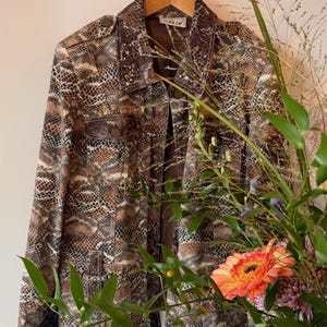 May include: A long-sleeve jacket featuring a brown, black, and white snakeskin pattern. The jacket includes a collared neckline, two chest pockets with button closures, shoulder epaulets, and small silver-toned studs along the collar and pockets.