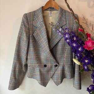 Può includere: Un blazer a quadri neri, bianchi e verde acqua è appeso a una gruccia di legno. Il blazer ha una chiusura a doppio petto con due bottoni neri. La fodera è color beige chiaro. Un mazzo di fiori viola e rosa è a destra.