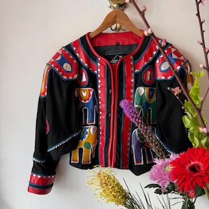 Peut inclure: Veste noire ornée d'éléphants colorés. La veste présente des bordures rouges, bleues et jaunes. Elle est suspendue à un cintre en bois. Des fleurs sont au premier plan. La veste mesure environ 50 cm de long.