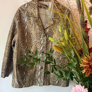 Könnte beinhalten: Eine langärmelige Jacke mit einem braun-beigen Schlangenledermuster. Die Jacke hat einen Reverskragen, eine Knopfleiste mit drei sichtbaren dunklen Knöpfen und ein helles Innenfutter, präsentiert auf einem Holzkleiderbügel.