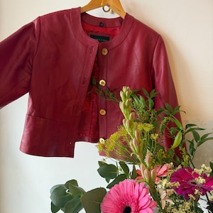 Puede incluir: Una chaqueta de cuero roja con cuello redondo y botones dorados cuelga de una percha de madera. La chaqueta se combina con un ramo de flores rosas y amarillas. El forro interior de la chaqueta es visible.