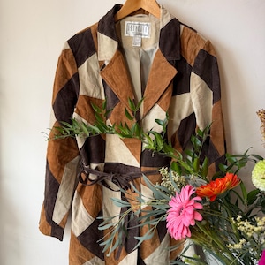 Puede incluir: Un abrigo vintage de ante patchwork en tonos marrón, beige y crema cuelga de una percha de madera. El abrigo presenta un patrón geométrico y un cinturón a juego. El abrigo se exhibe contra una pared neutra.