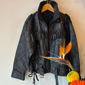 Könnte beinhalten: Eine schwarze Lederjacke mit Fransen, goldenen Stern-Applikationen und einem Schnürdetail an der Seite. Die Jacke hat einen Reißverschluss und einen Gürtel.