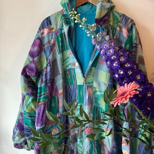 Op de afbeelding: Een kleurrijke patchworkjas met capuchon en ritssluiting. De jas heeft een levendige mix van paarse, groene en blauwe tinten. De binnenvoering is turquoise. De jas hangt aan een houten kleerhanger.