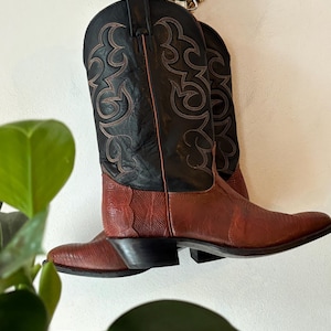 Op de afbeelding: Een paar bruine leren cowboylaarzen met zwarte leren accenten en ingewikkelde stiksels. De laarzen hebben een spitse neus en een lage hak.