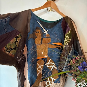 Op de afbeelding: Een bruin, blauw en beige patchworkjack met een figuur van een krijger die een zwaard in het midden vasthoudt. Het jack heeft een ronde hals en lange mouwen.