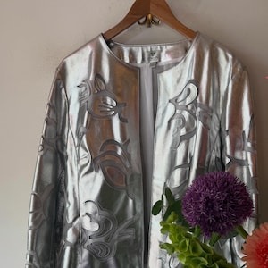 Op de afbeelding: Zilveren jas met een bloemenpatroon. De jas hangt aan een houten hanger. De jas heeft een glanzende, metallic afwerking. De jas is open en onthult een witte voering. Een boeket bloemen staat op de voorgrond.