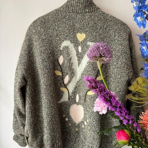 Op de afbeelding: Een groene gebreide trui met een hoge kraag, met een wit bloemmotief met roze en gele accenten. De trui hangt aan een houten hanger. Een boeket paarse en roze bloemen staat op de voorgrond.