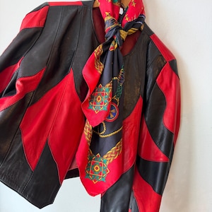 Op de afbeelding: Een vintage leren jas met een opvallend colorblock-ontwerp in zwart en rood. Een zijden sjaal met patronen in rood, zwart en goud hangt over de jas. De jas hangt aan een houten kleerhanger.