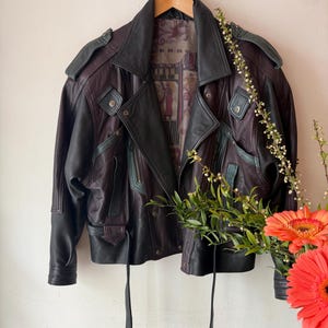 Könnte beinhalten: Eine Vintage-Lederjacke in Burgunderrot und Schwarz hängt an einem Holzbügel. Die Jacke hat einen klassischen Biker-Stil mit einem Reverskragen, Schulterklappen und mehreren Taschen. Das Innenfutter hat ein Muster.