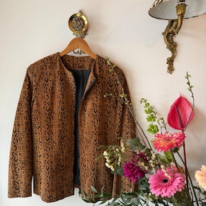 Puede incluir: Una chaqueta marrón y negra con estampado de leopardo y forro negro cuelga de una percha de madera. La chaqueta está con cremallera y tiene mangas largas. Se ve un aplique de pared vintage con una pantalla de lámpara con estampado de leopardo.