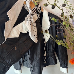May include: Black leather jacket with white detailing and matching trousers. A polka dot scarf and a monochrome patterned scarf hang on the hanger. The jacket features a zip and a quilted design.