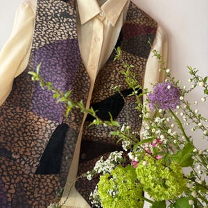 Può includere: Un gilet patchwork vintage con chiusura a bottoni, caratterizzato da un mix di tessuti fantasia nei toni del marrone, viola e nero. Il gilet è sovrapposto a una camicia con colletto color crema. Un bouquet di fiori nei toni del verde, viola e bianco è in primo piano.