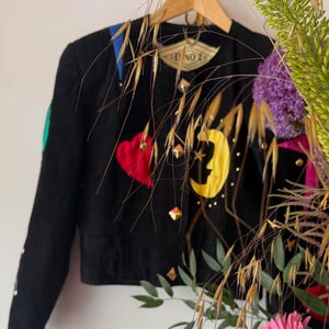 Op de afbeelding: Een zwarte jas met een rode hartapplicatie, een gele halve maan met een ster en goudkleurige piramidevormige studs. Het kledingstuk heeft een ronde halslijn, lange mouwen en een label in de kraag met de tekst D NO Z op een hanger.