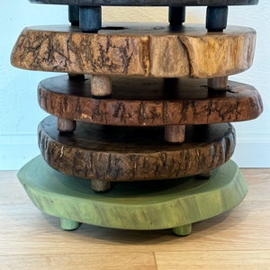 May include: A unique, handcrafted wooden pedestal with four stacked, circular platforms. The platforms are various colors, including black, natural wood, and green, and are supported by short, cylindrical legs. The wood grain is visible.