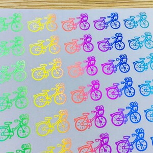 May include: Sheet of iridescent bicycle stickers in various colors, including green, pink, and blue. The stickers are arranged in rows and columns on a white background. A black ruler, pens, and panda-shaped decorations are also visible.