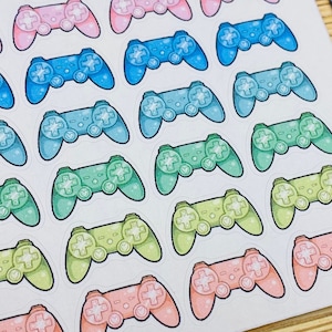 May include: A sheet of colorful stickers featuring video game controllers in pastel shades of pink, blue, green, and yellow. The controllers are arranged in rows on a white background. A black ruler, scissors, and a panda figurine are also visible.