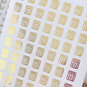 May include: A sheet of gold foil stickers with a calculator design. The stickers are on a white background and are arranged in a grid pattern.