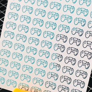May include: A sheet of stickers with a repeating pattern of blue and black video game controllers. The controllers are outlined in black and have a gradient of blue to black.