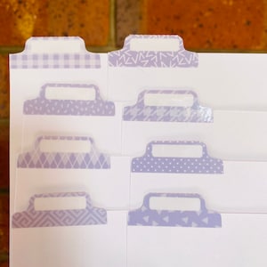 May include: Eight purple and white patterned index tabs with blank white labels. The tabs have a variety of patterns including gingham, geometric, and polka dots.