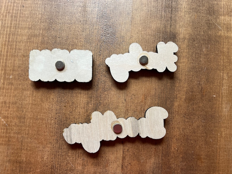 May include: Three wooden signs with the words "Home", "Welcome", and "Blessed" written on them. Each sign has a small black magnet attached to the back.