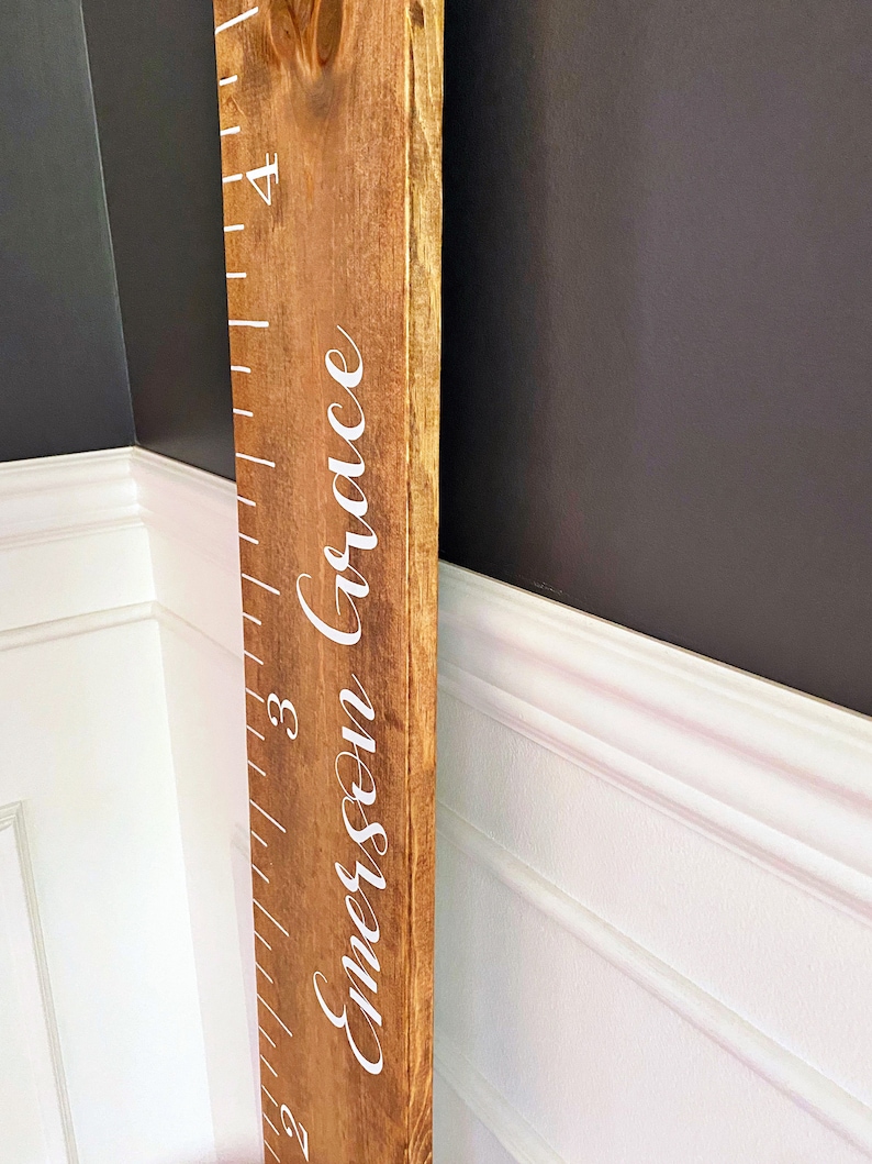 May include: A wooden growth chart with a ruler and the name "Emerson Grace" written in white cursive lettering.