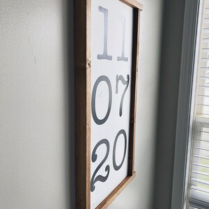 K&ouml;nnte beinhalten: Ein gerahmtes Holzschild mit dem Datum "11 07 20" in Schwarz auf wei&szlig;em Hintergrund.