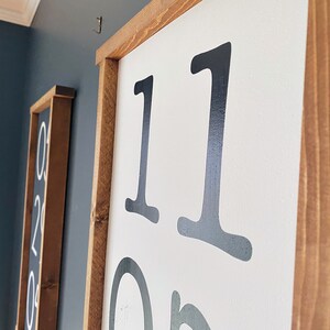 K&ouml;nnte beinhalten: Nahaufnahme eines Holzschildes mit der Nummer "11" in schwarzer Schrift auf wei&szlig;em Hintergrund. Das Schild h&auml;ngt an einer Wand.
