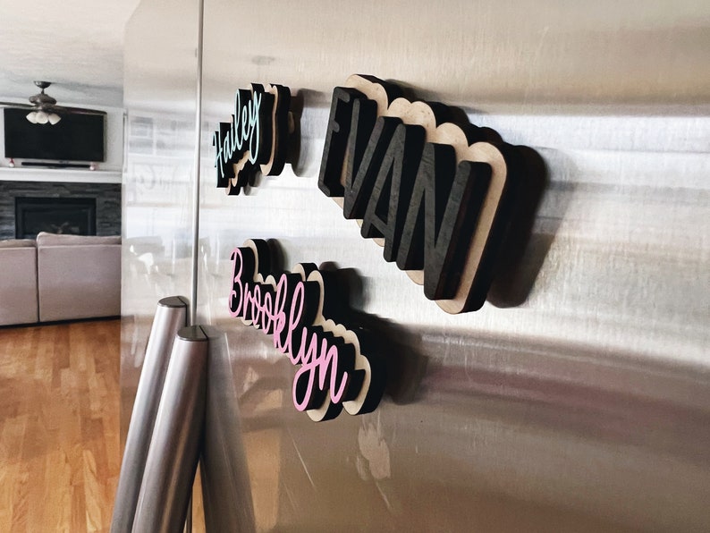 May include: Four wooden name signs with a 3D effect, attached to a stainless steel refrigerator. The names are "Hailey", "Evan", "Brooklyn", and "Brooklyn". The names are painted in different colors, including pink, teal, and black.