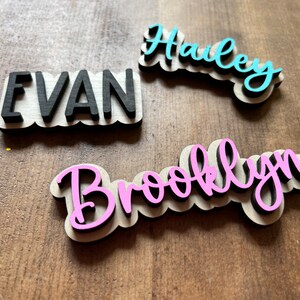 May include: Three wooden name plaques with names "Evan", "Hailey", and "Brooklyn" written in black, blue, and pink paint, respectively. The plaques are on a wooden surface.